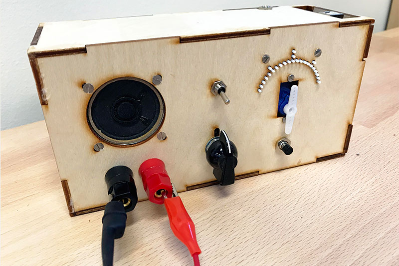 A wooden box which houses the hardware for the accessible voltmeter. The front has buttons, tactile marks, lead probes, and  a speaker panel.