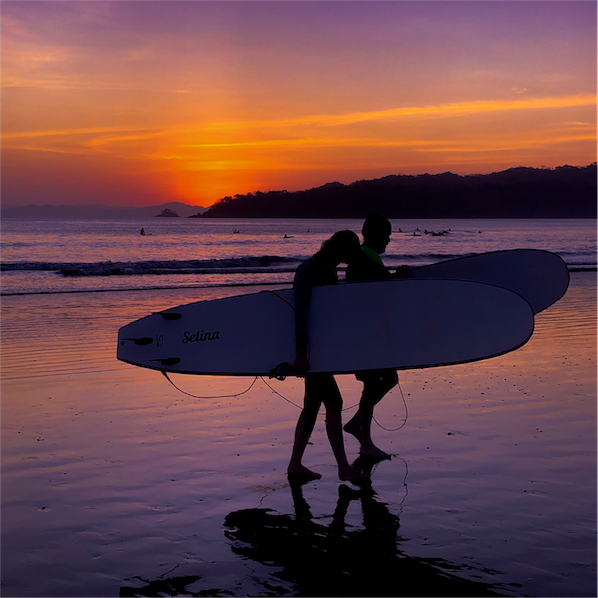 Sunset facing the ocean making the water and sky colors have orange and pink hues. In the foreground there is the silhouette of a couple each carrying surfboards.